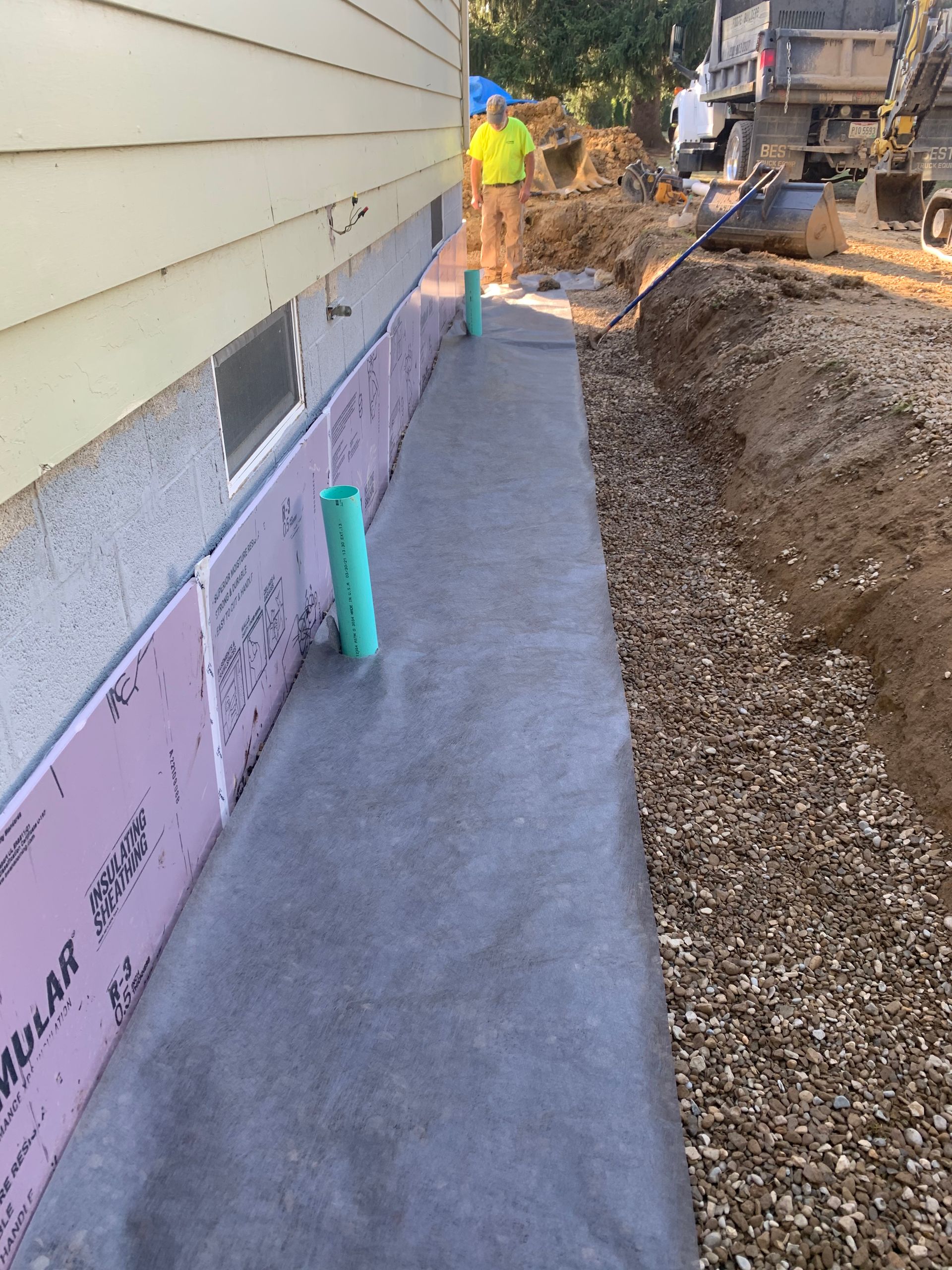 Foundation work next to a light yellow house. Pink insulation, gray fabric, and gravel line a trench.