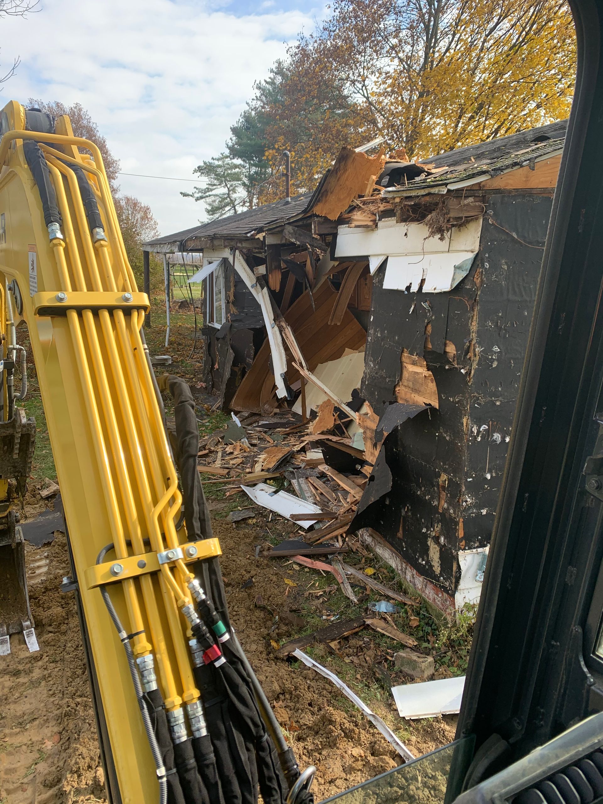 Excavator demolishing a damaged house; exterior walls and roof partially collapsed, wooded background.