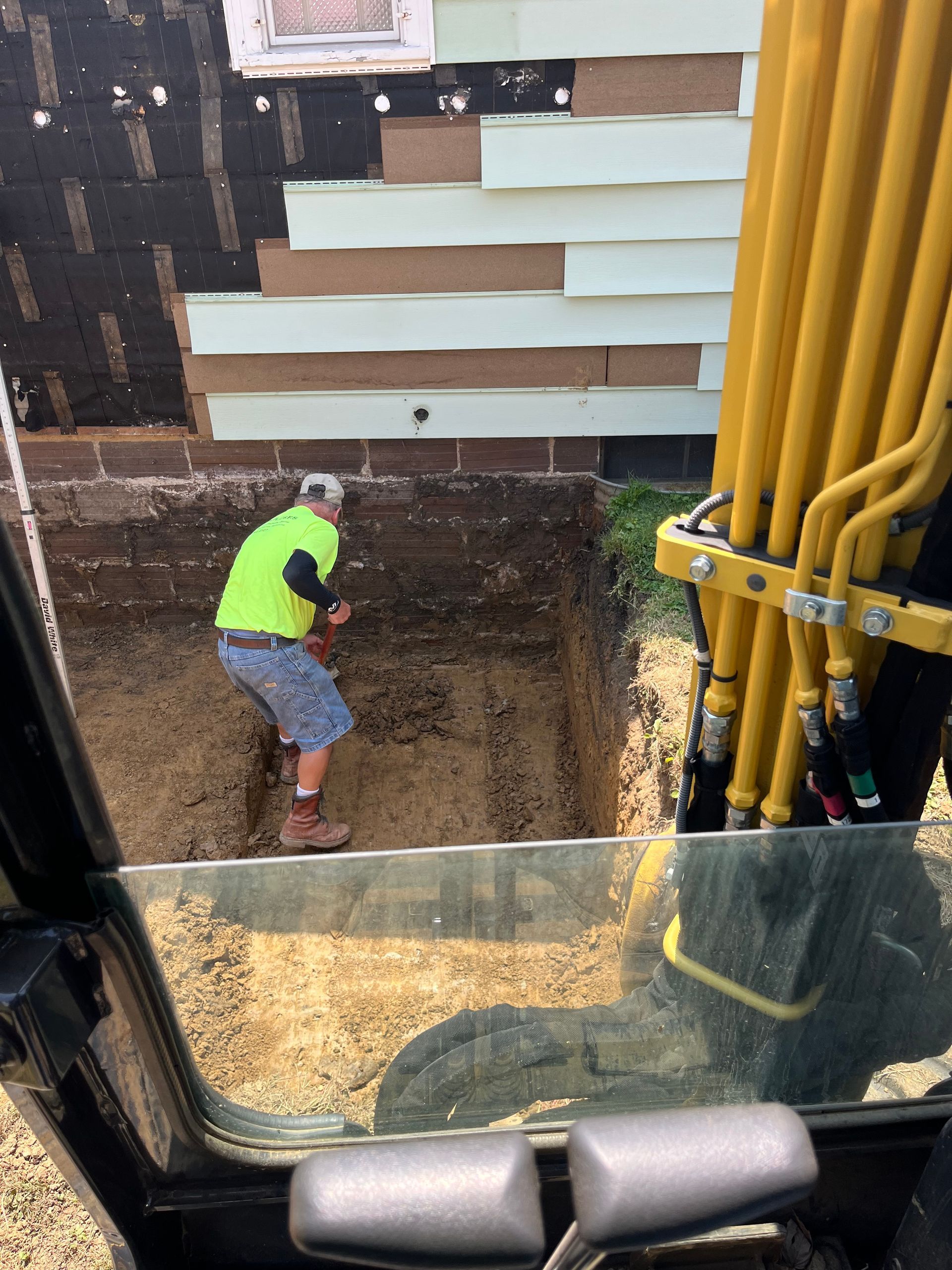 Man in safety vest digging near foundation of a building. Yellow excavator arm is visible.