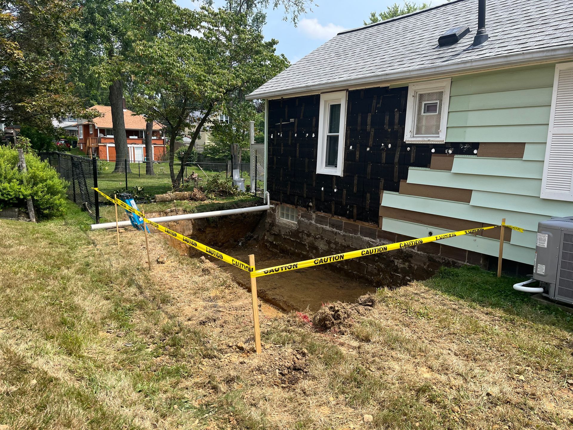 Excavation beside a house with exposed foundation and caution tape. The siding is partially removed.