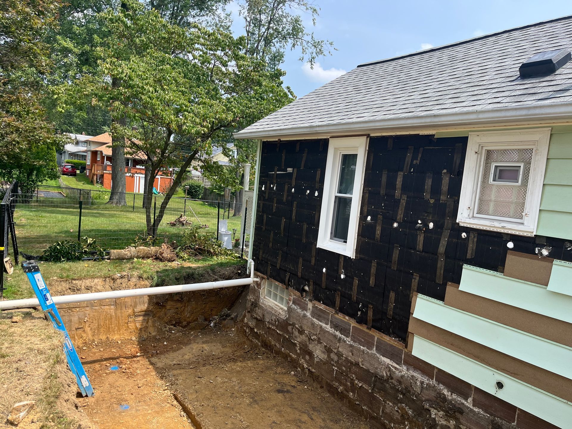 Exterior house under renovation, side view. Siding removed, revealing black underlayment. Excavated area in front.