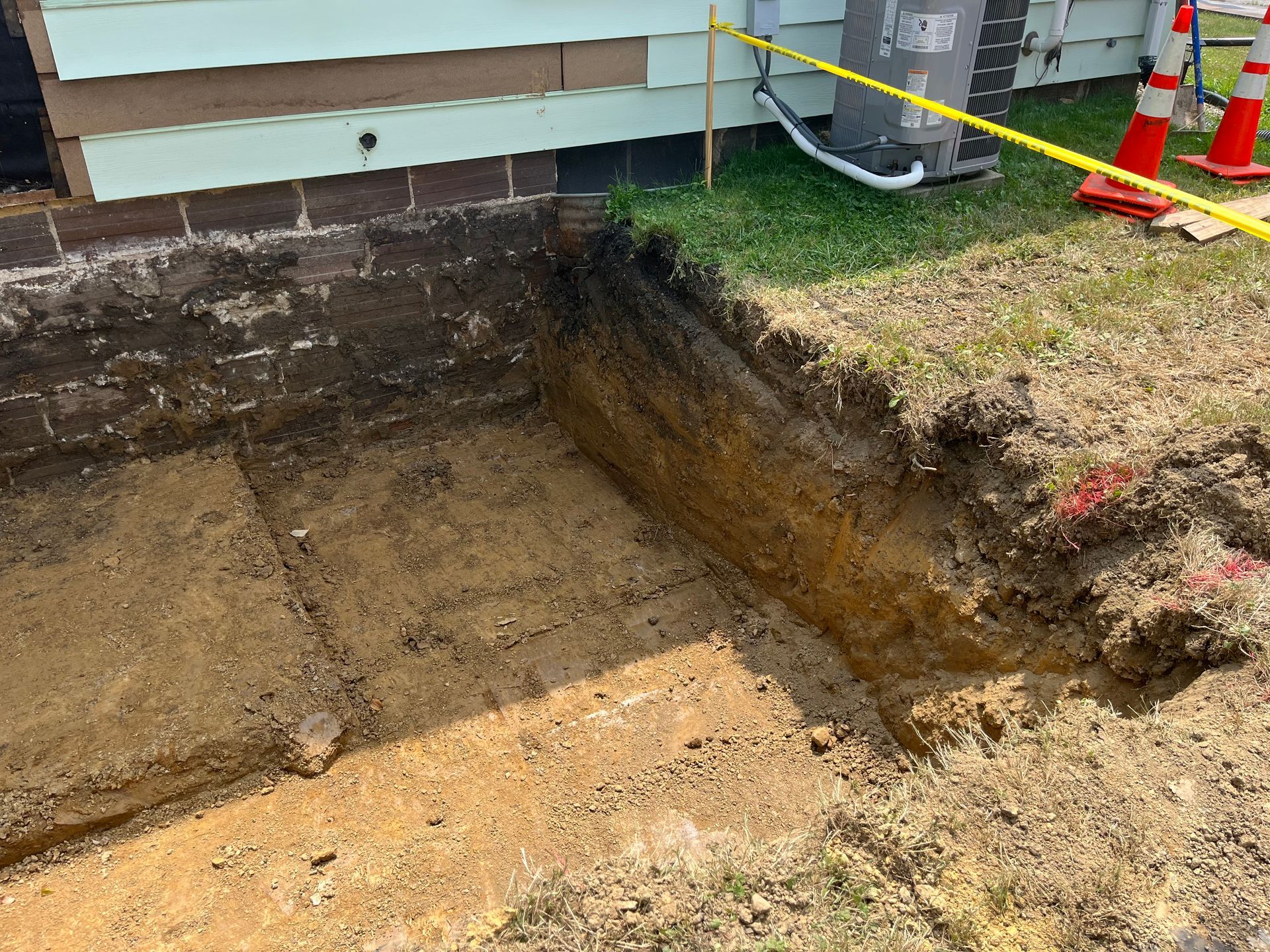 Excavation next to a house with exposed foundation and soil layers. Yellow caution tape and orange cones in grass.