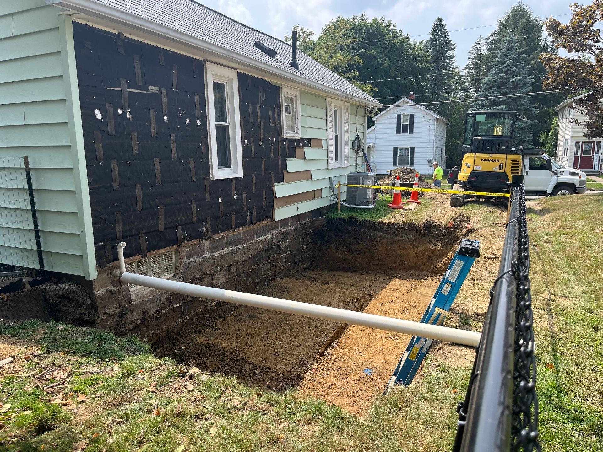 House exterior with excavation in progress, exposing the foundation, white pipe, and a small excavator.
