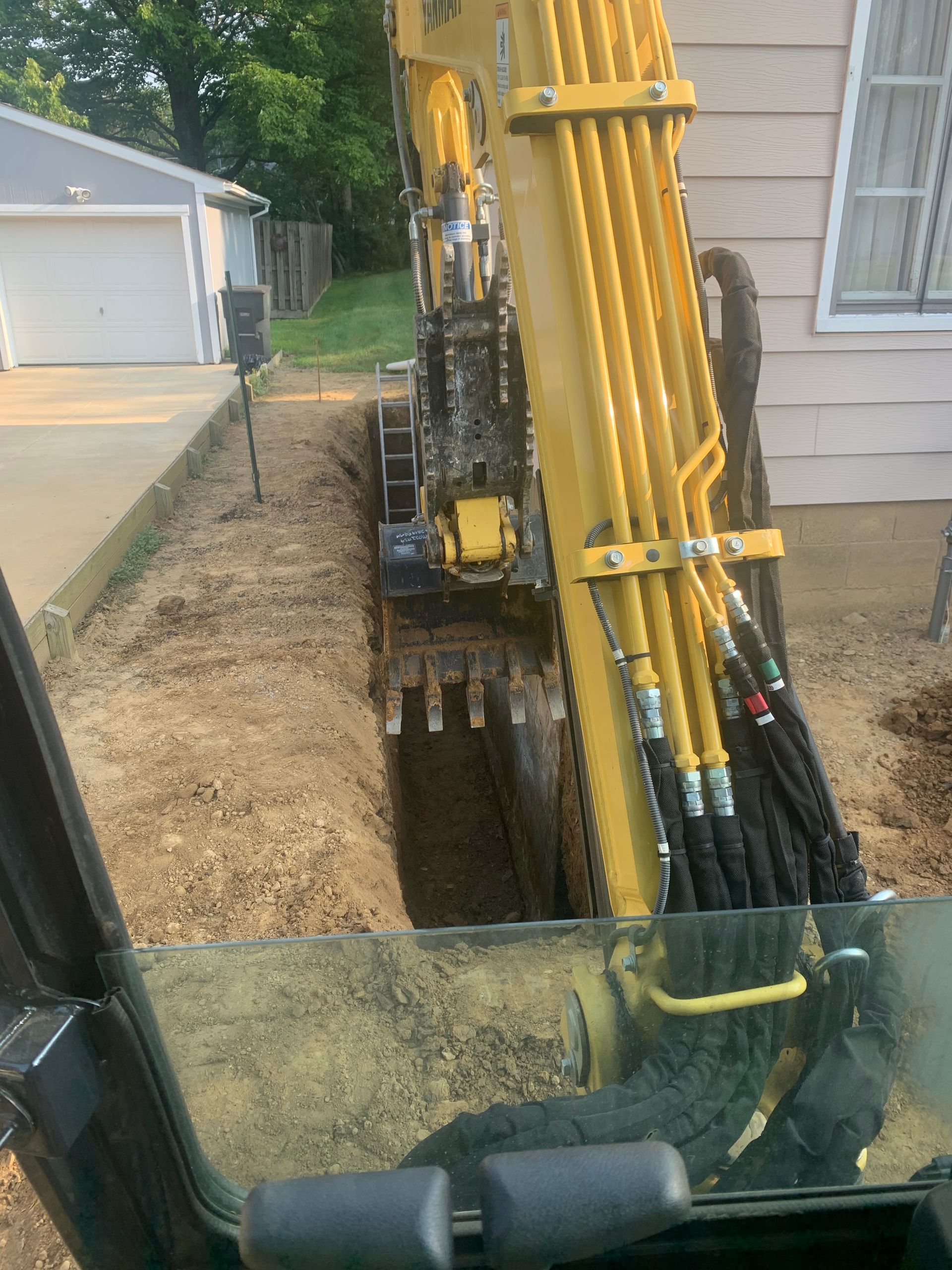 A yellow excavator digging a trench next to a driveway and house.