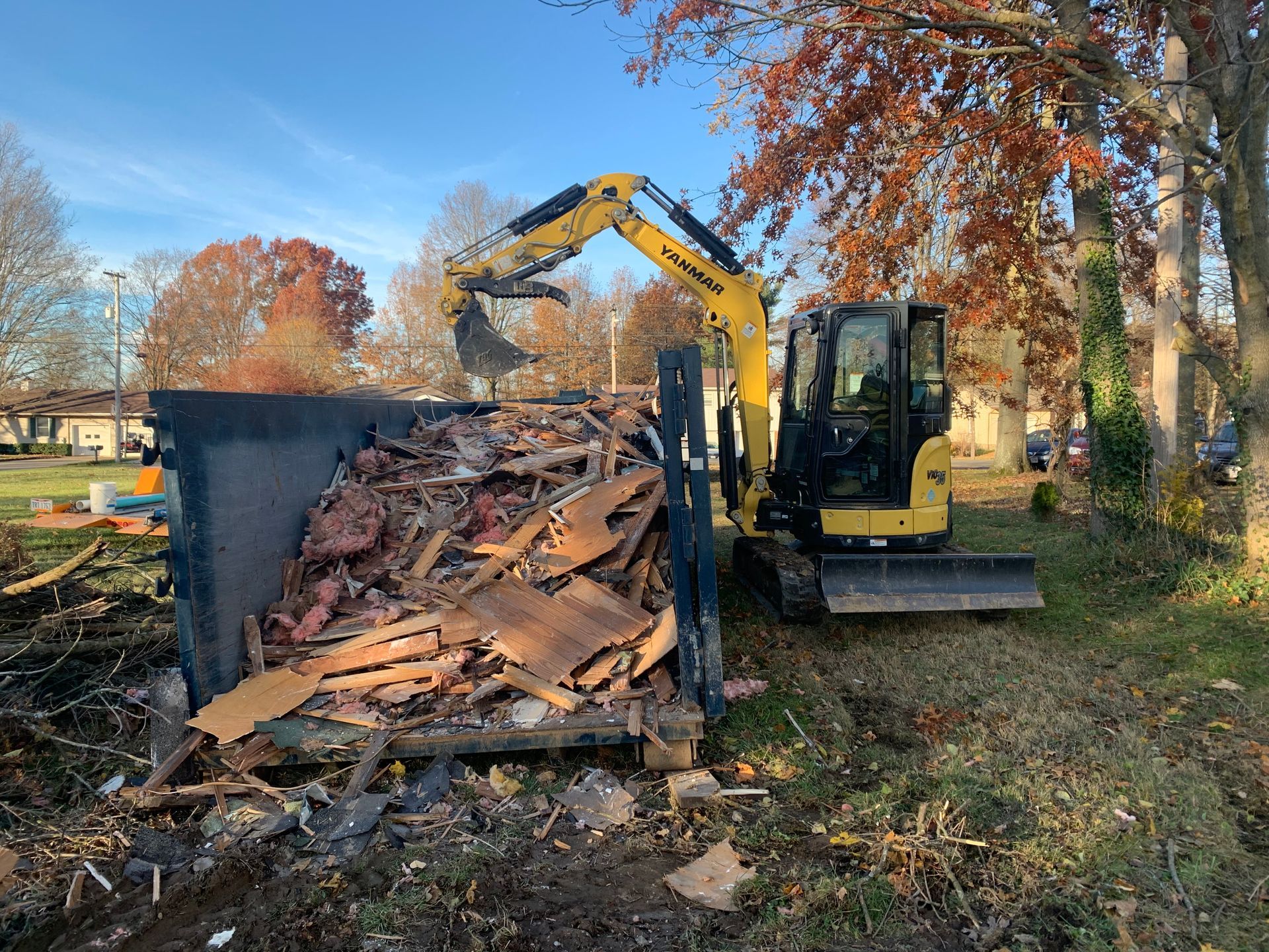 Excavator loads debris into a dumpster outdoors with fall foliage.