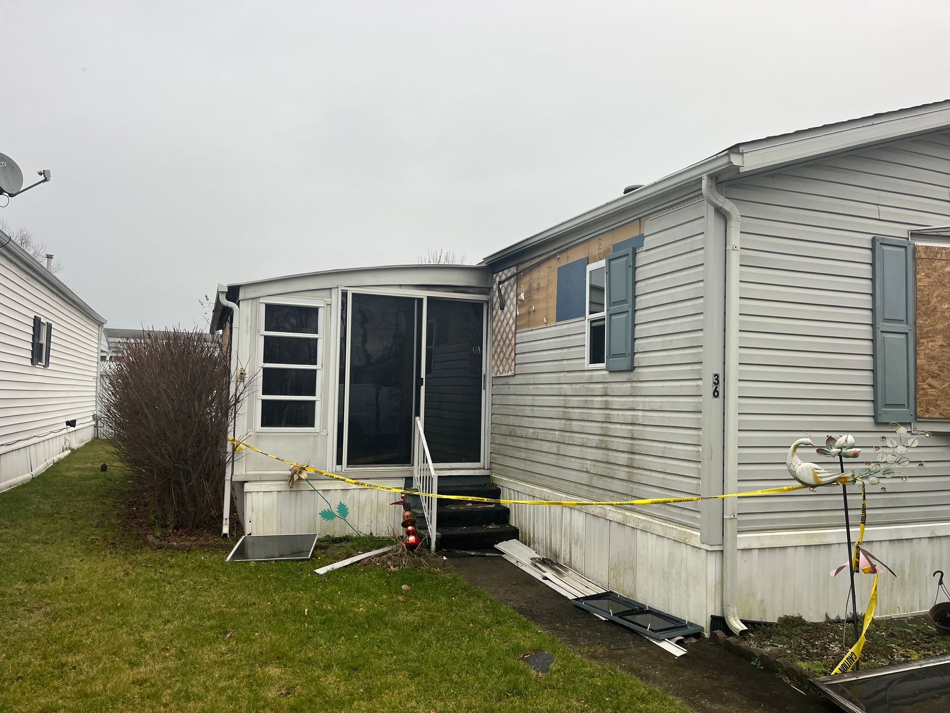 Damaged mobile home exterior with boarded windows, surrounded by caution tape, cloudy day.