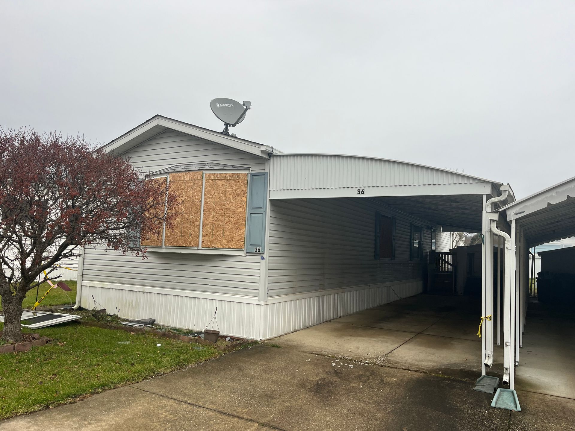 Mobile home with boarded-up window, carport, satellite dish, and overcast sky.