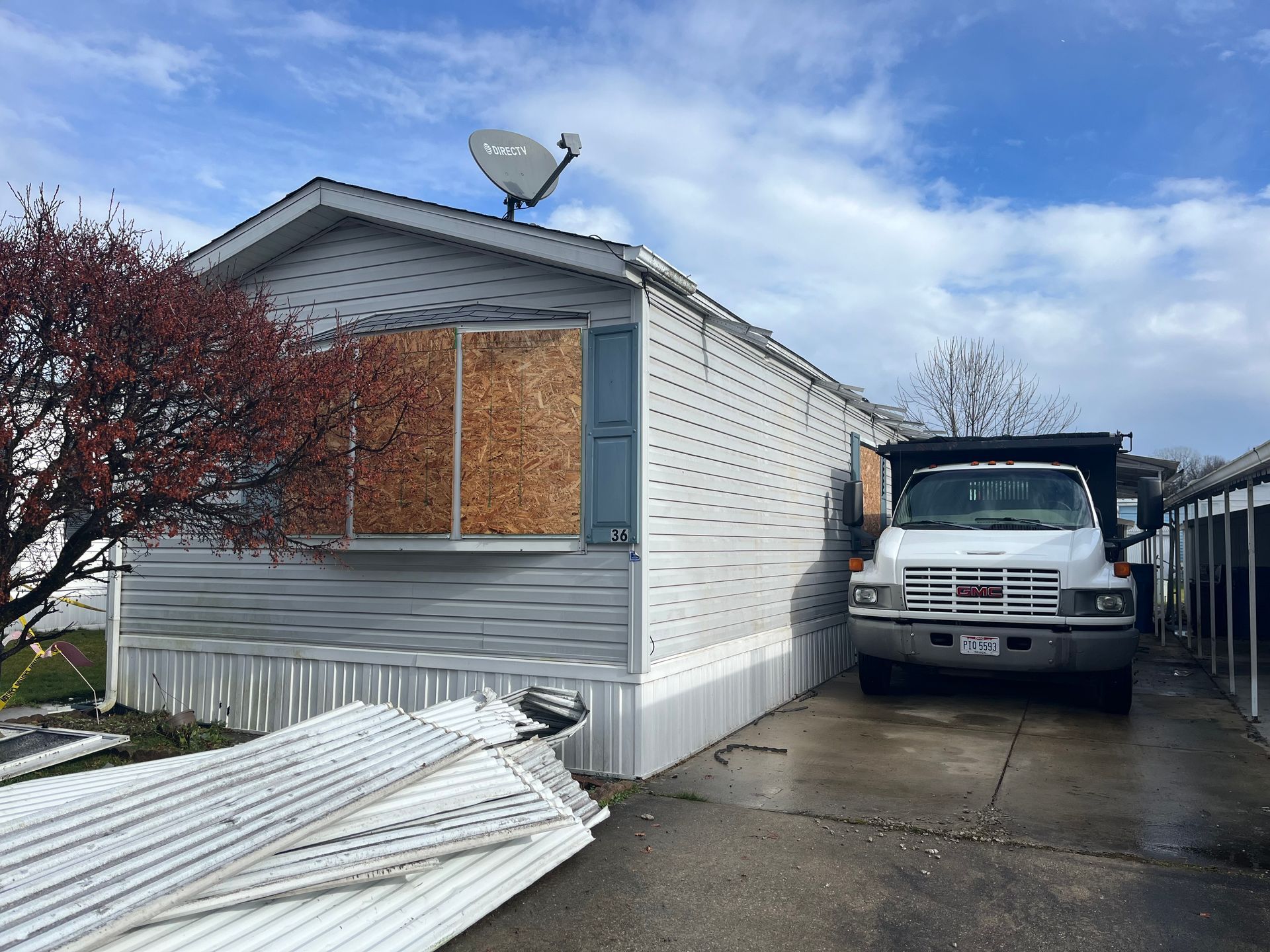 Mobile home with boarded-up windows; truck parked in the driveway. White siding, satellite dish, and cloudy sky.