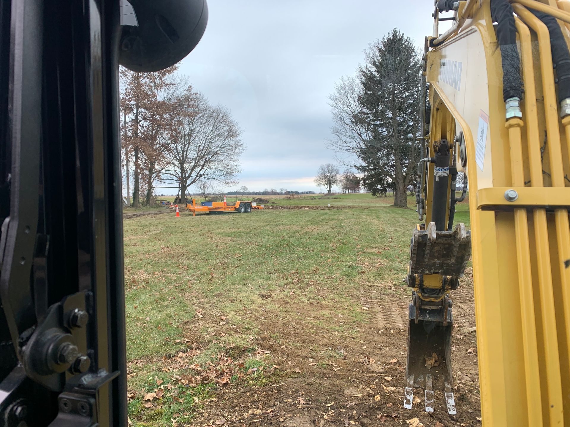 View from excavator cab with a construction site; field, trees, and other machinery visible.