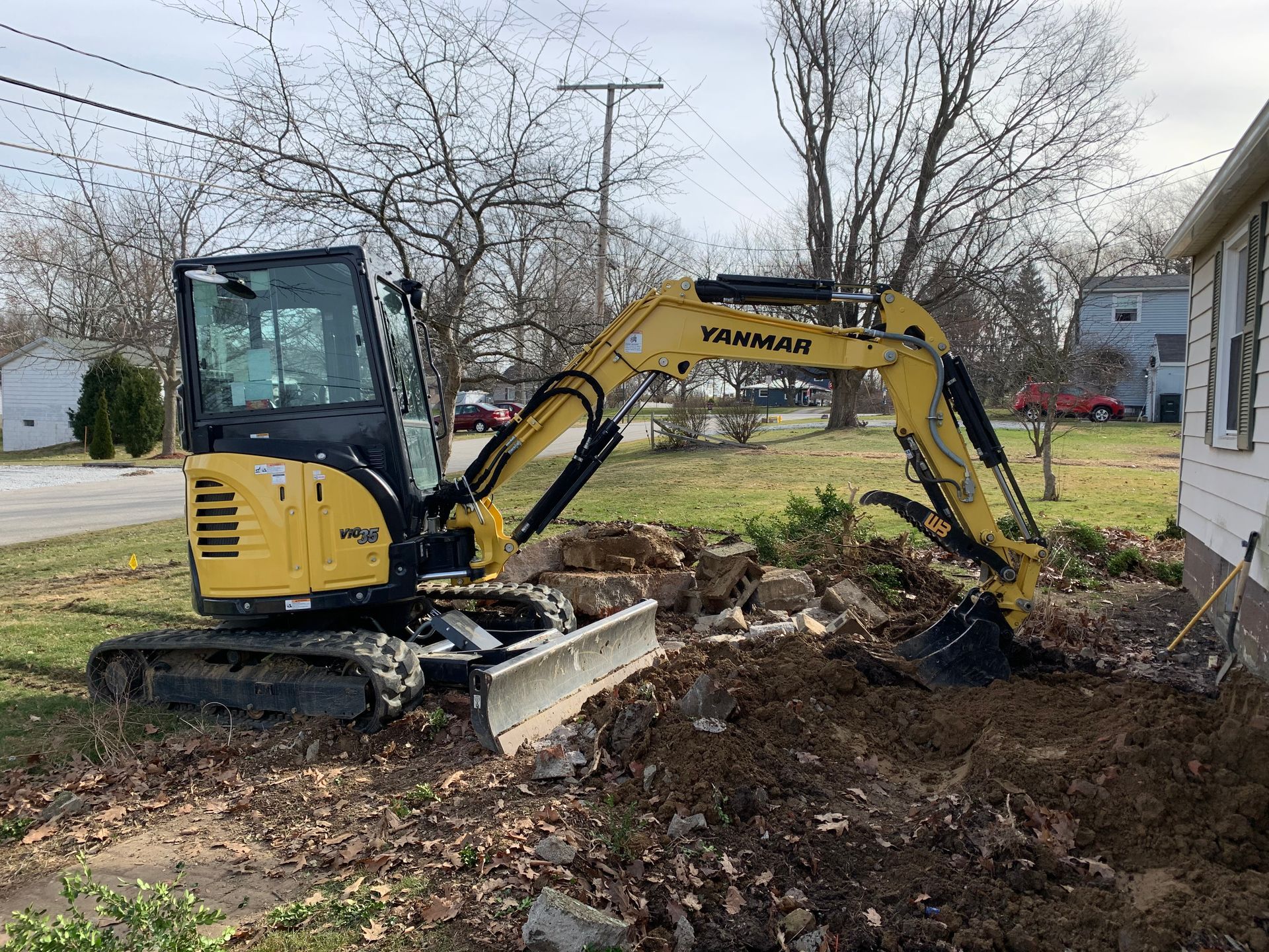 Yellow Yanmar mini excavator digging in a yard next to a house.