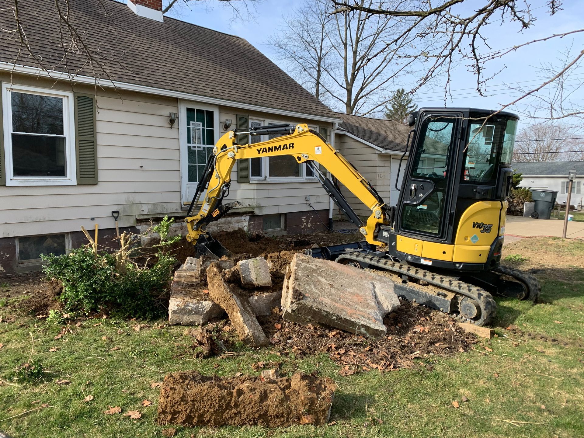 Yellow excavator digging near a house, debris on the ground.