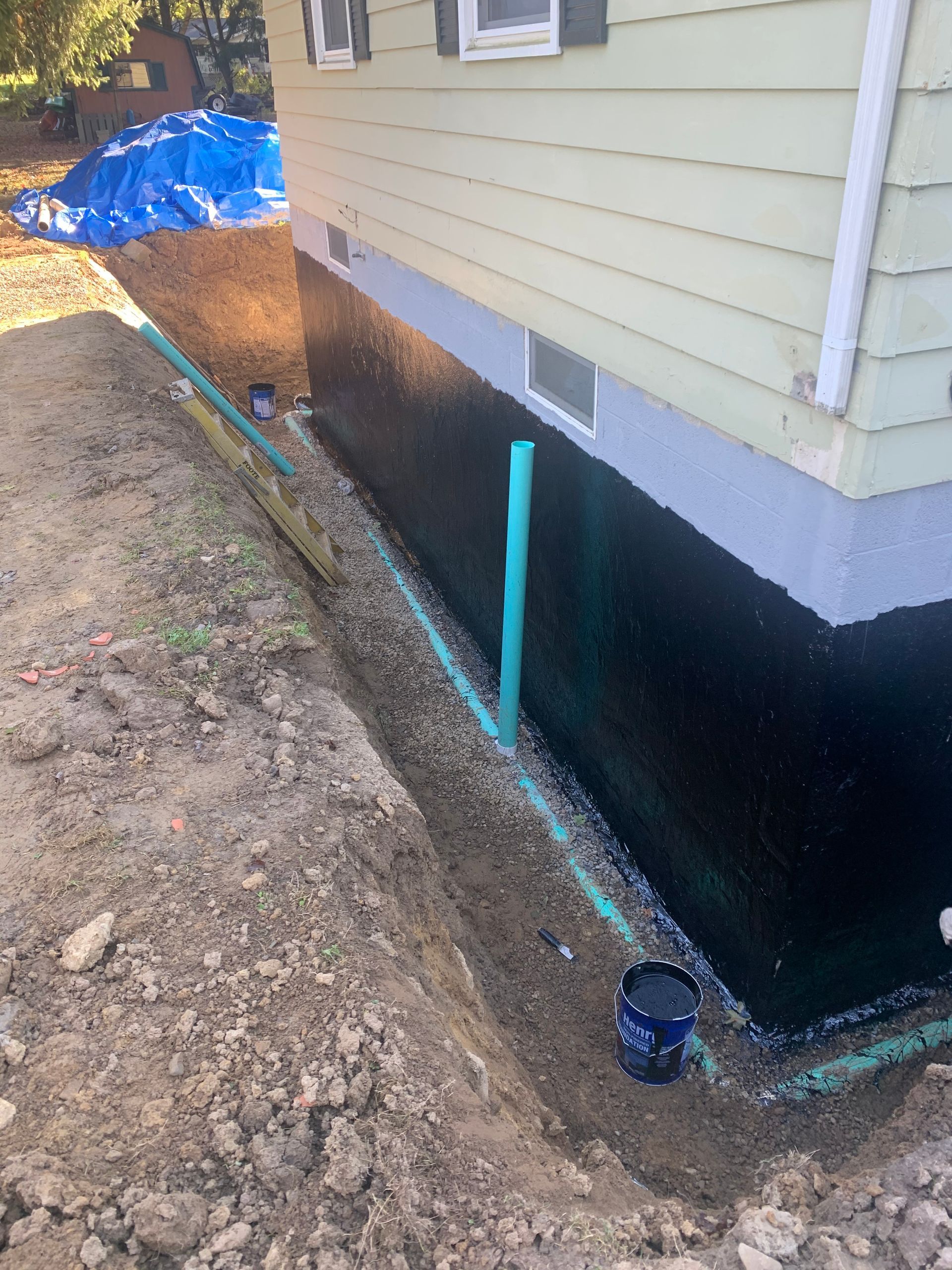 Exterior house foundation with applied black waterproofing and drainage pipe in a trench.