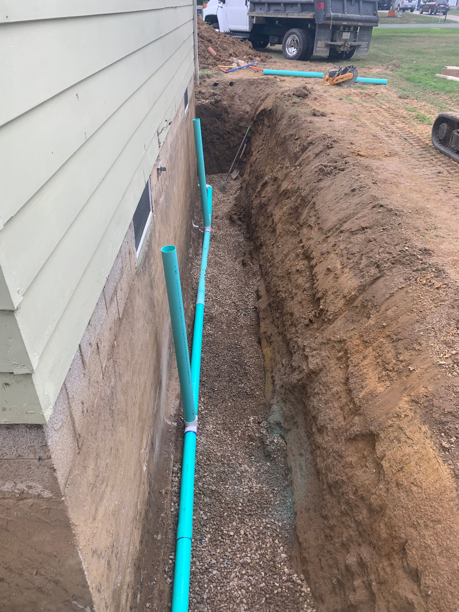 Trench next to a building with green pipes and gravel. Construction equipment visible in the background.
