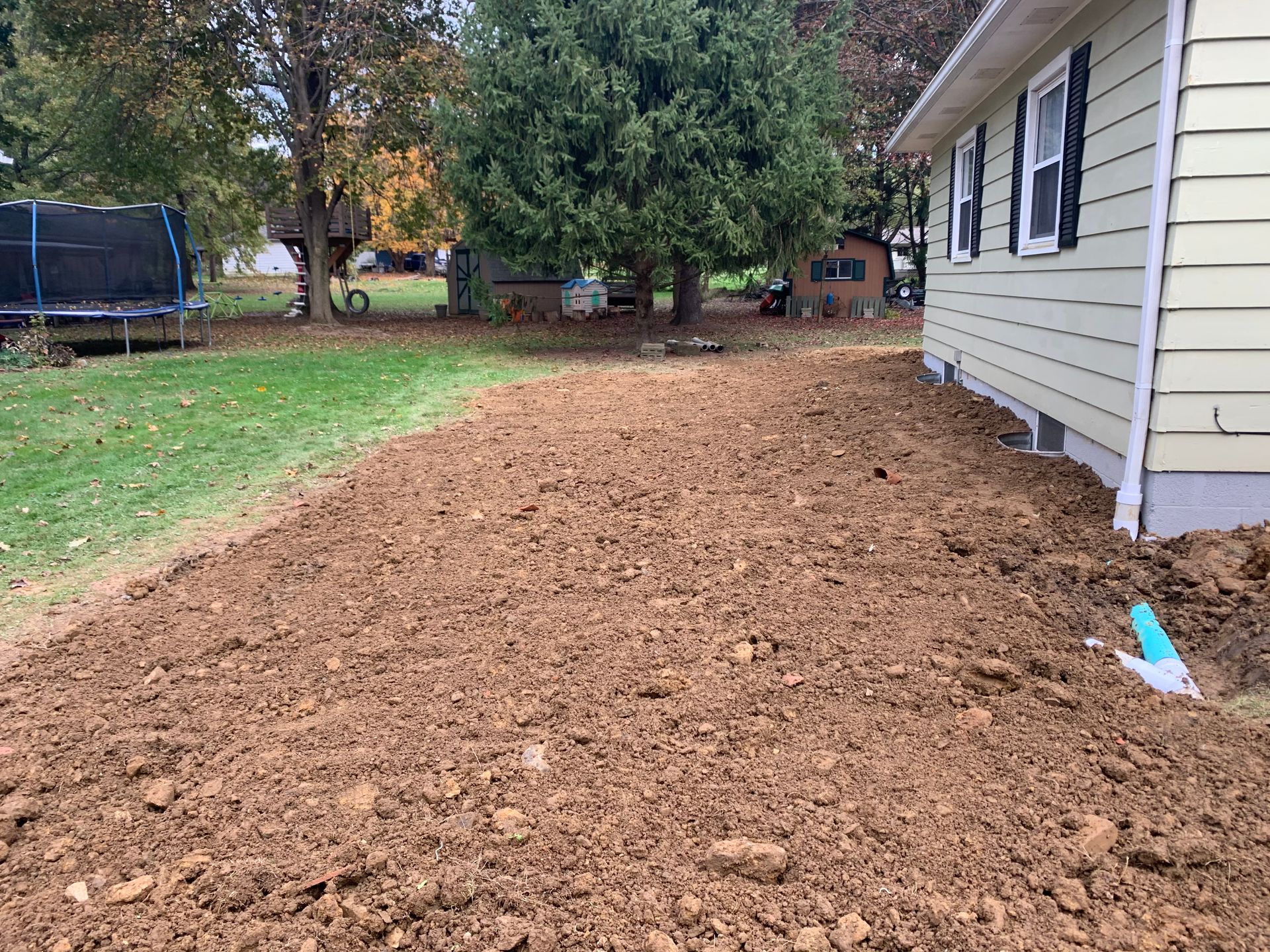 Tilled dirt beside a house with tan siding, a yard, and a trampoline in the background.