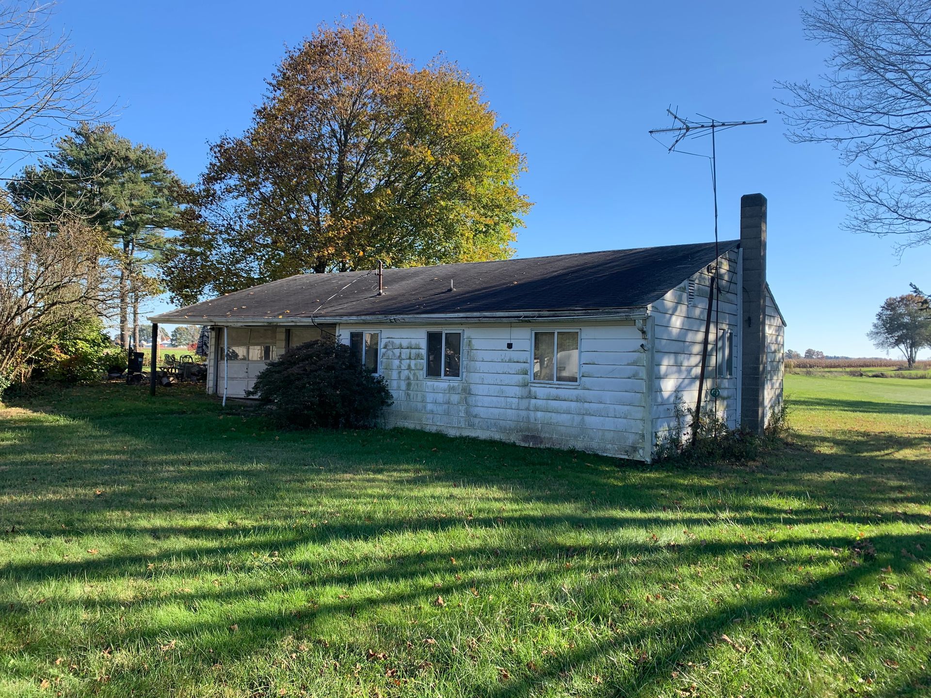 Old, white, single-story house with a chimney and antenna in a grassy field on a sunny day.