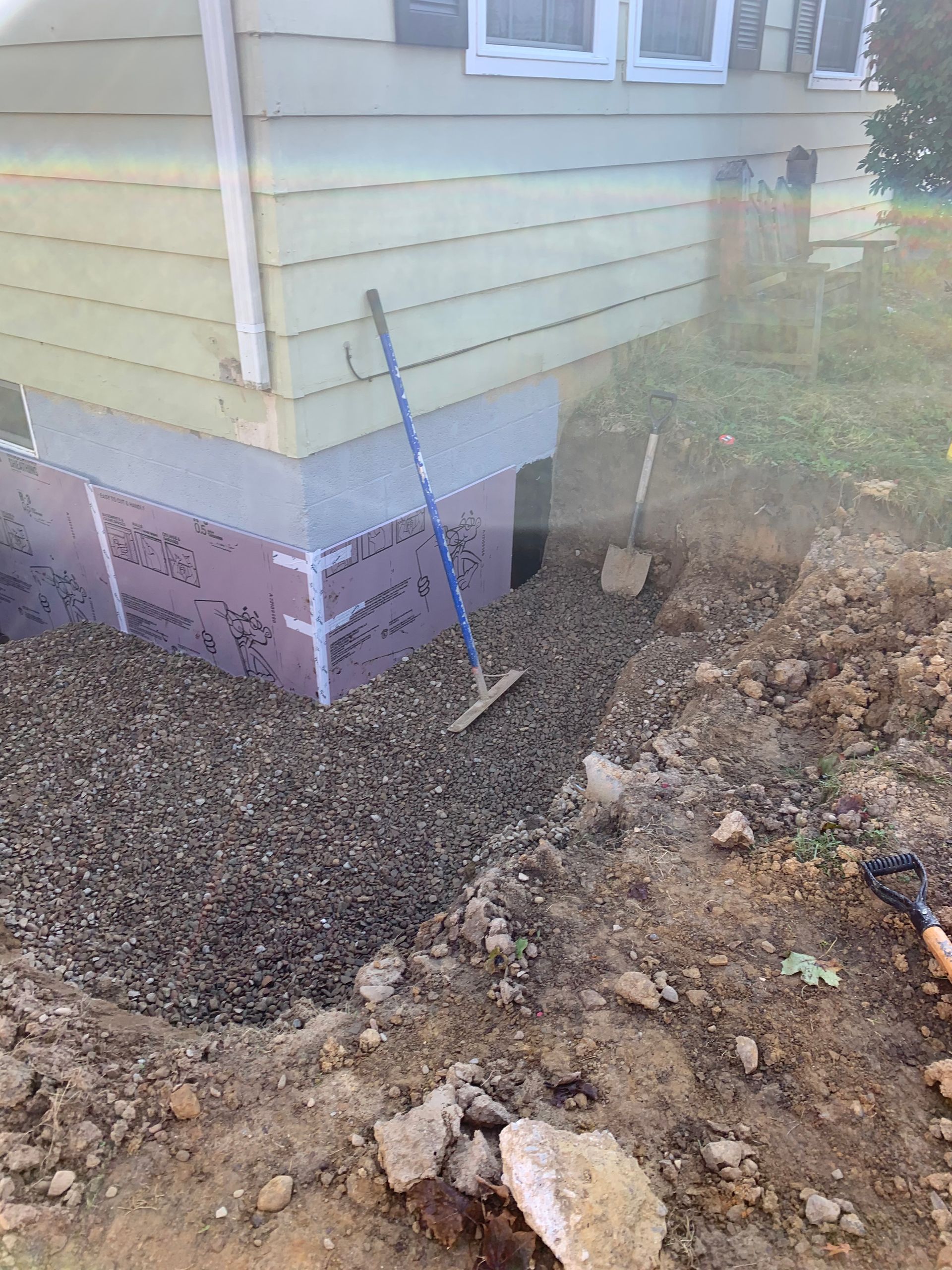 Excavation near a house foundation, showing gravel, insulation, and tools like a rake and shovel.