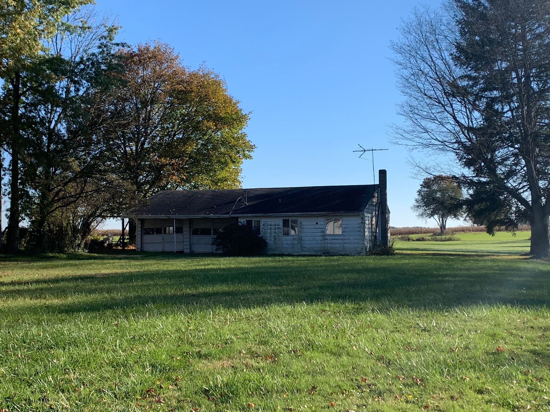 Dilapidated, blue-painted house with a dark roof and chimney, surrounded by green grass and trees, under a blue sky.