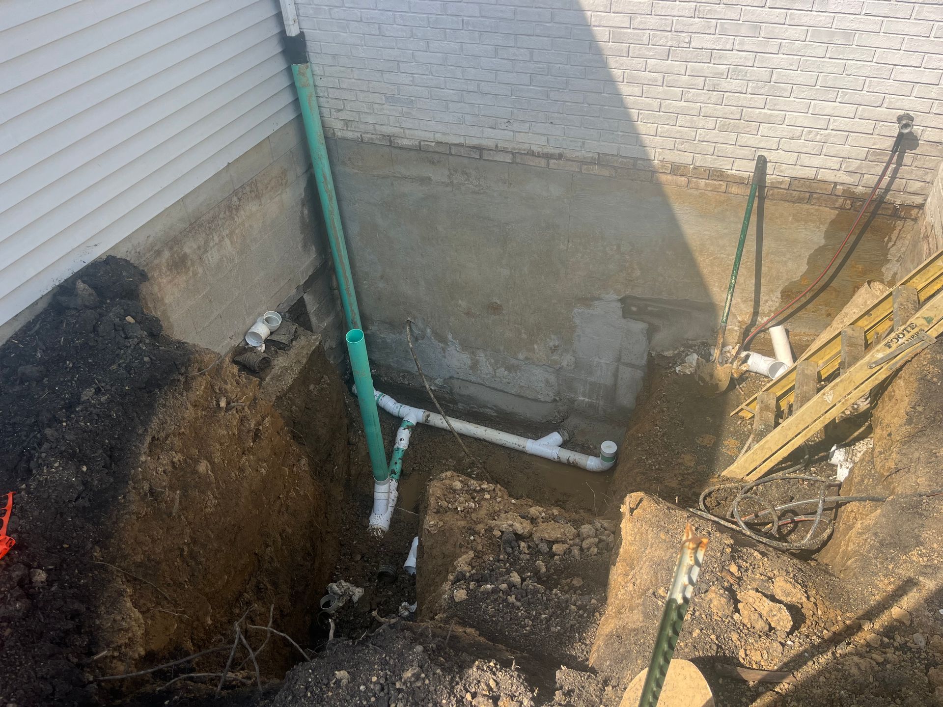 Excavation next to a building foundation with exposed plumbing and green pipes.