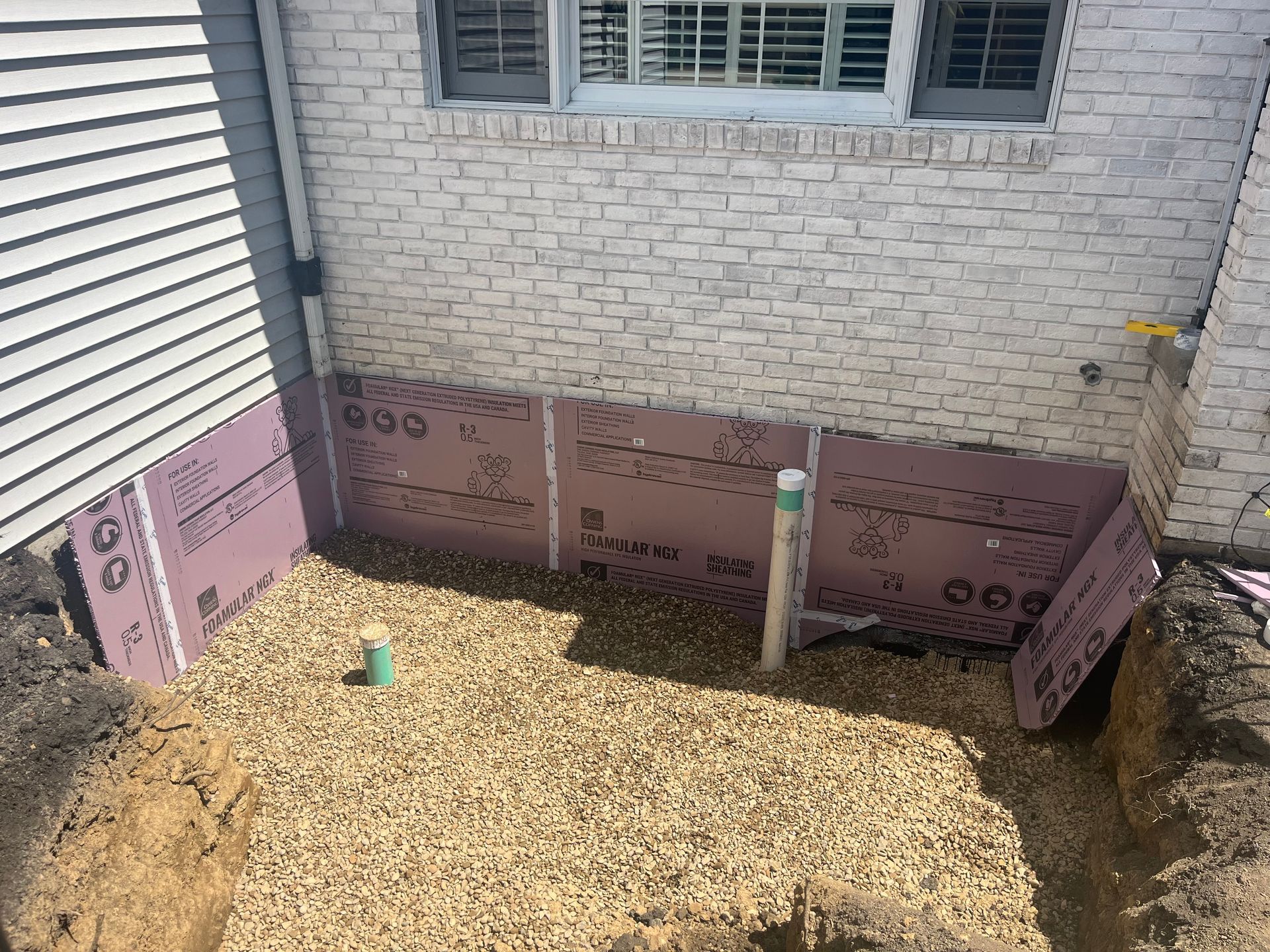 Construction site: pink foam insulation panels against a brick wall, gravel base, green pipe, exposed foundation.