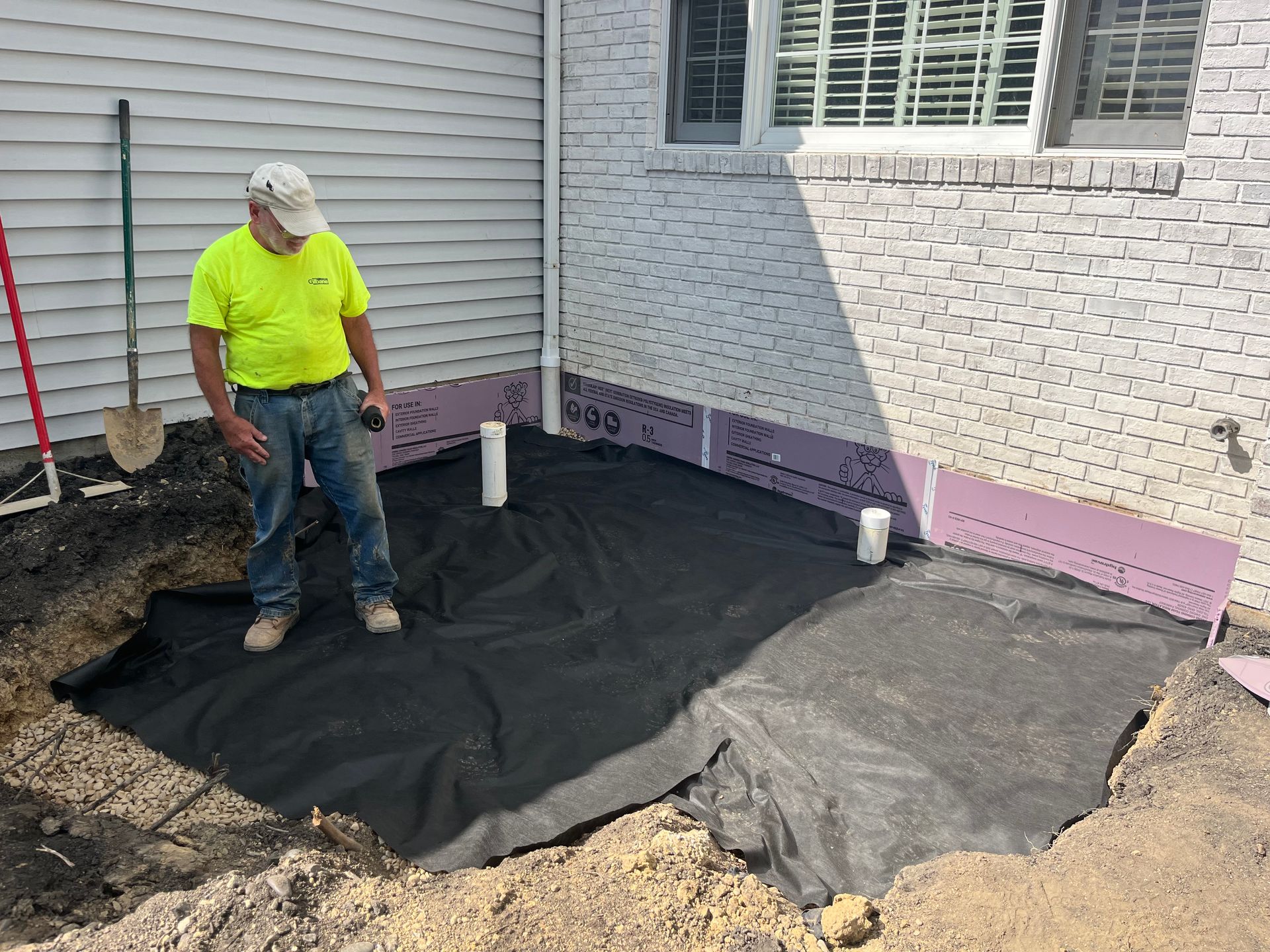 Man stands in a dug-out area with fabric, pipes, and insulation near a building's foundation.