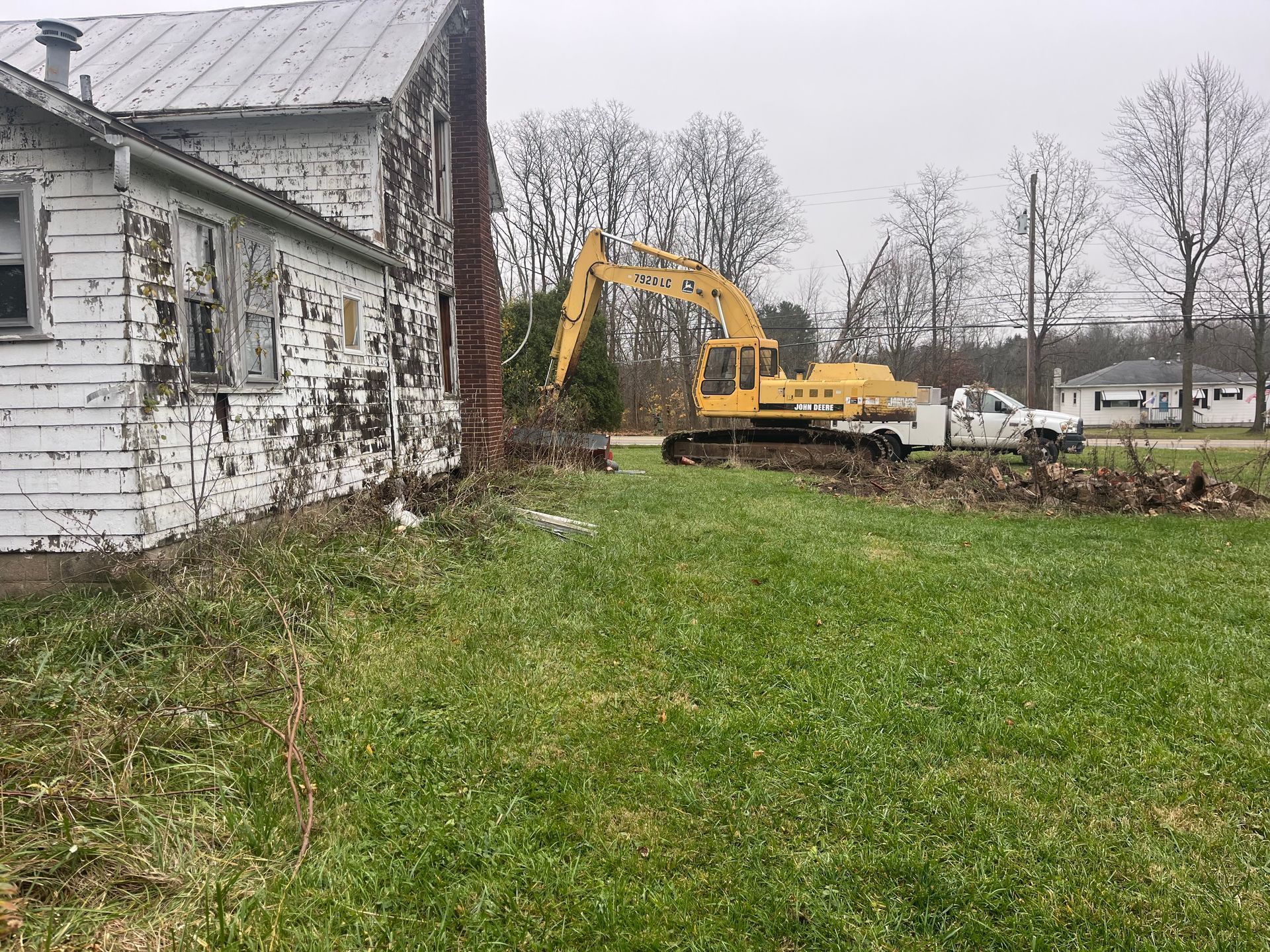 A yellow excavator is demolishing an old white house on a grassy lot.