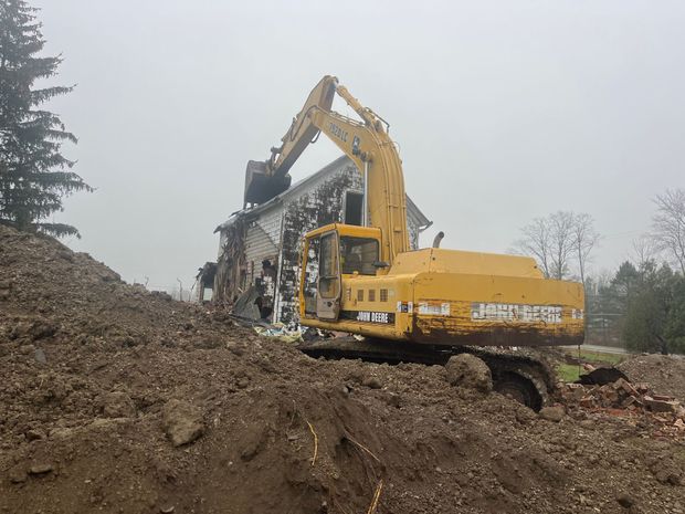 Yellow excavator demolishing a white house on a cloudy day. Debris and dirt surround the scene.