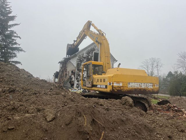 Yellow excavator demolishing a white house on a cloudy day. Debris and dirt surround the scene.