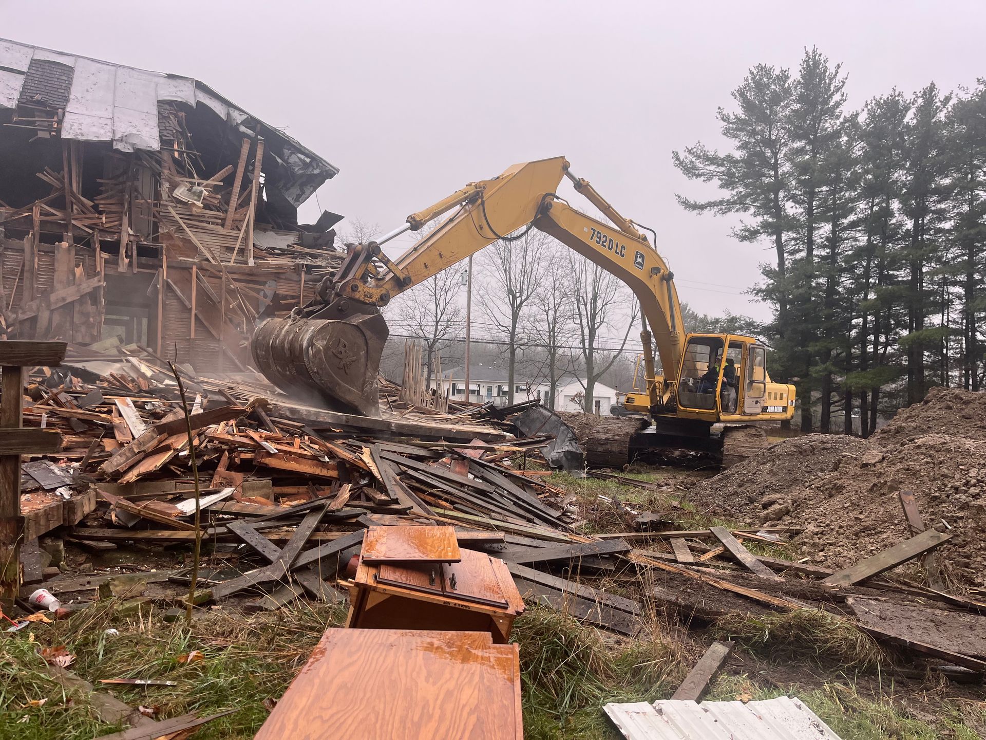 A yellow excavator demolishes a barn, surrounded by debris. Overcast day.