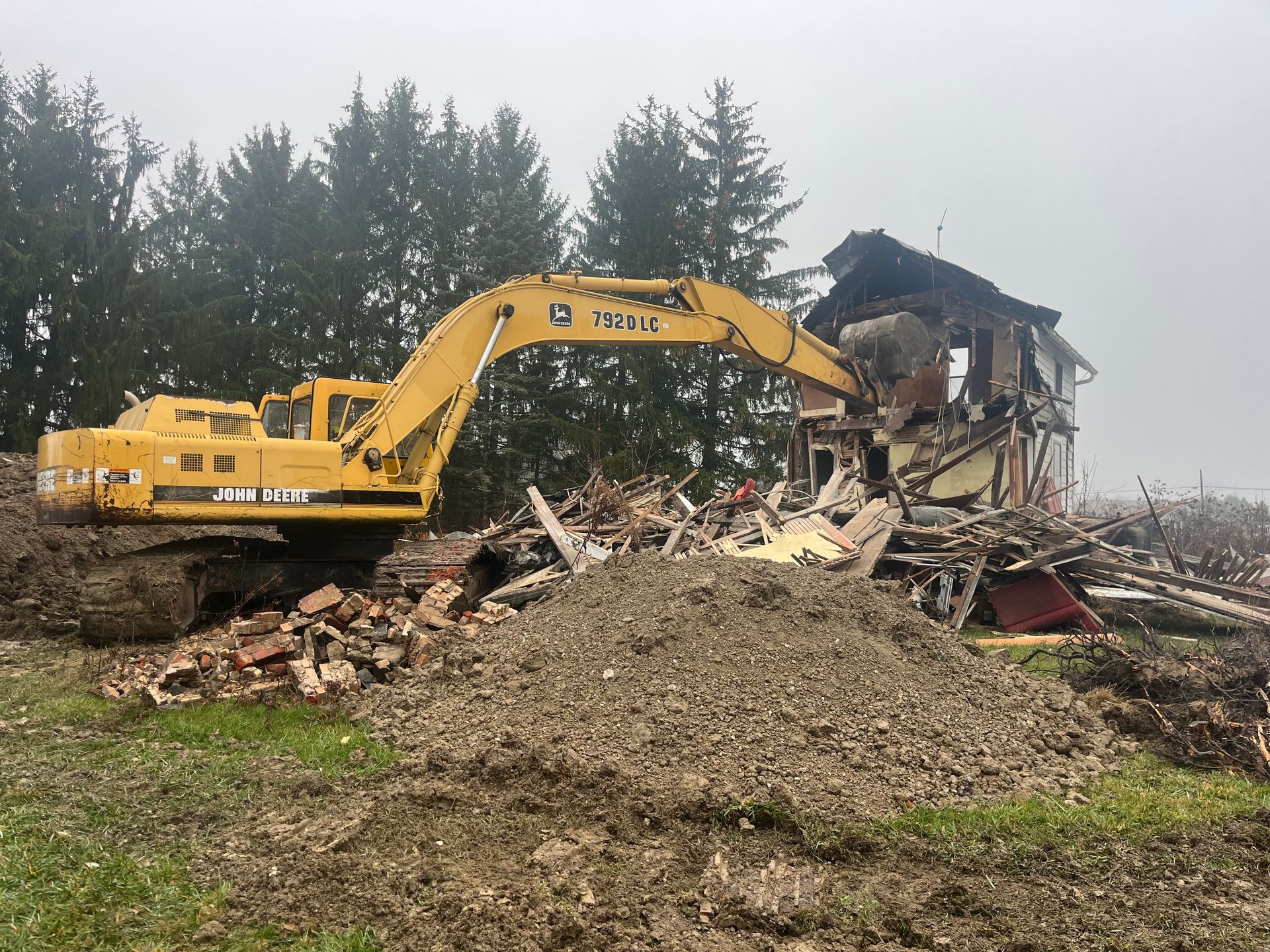 Yellow excavator demolishes a house; debris scattered on green grass, trees in the foggy background.