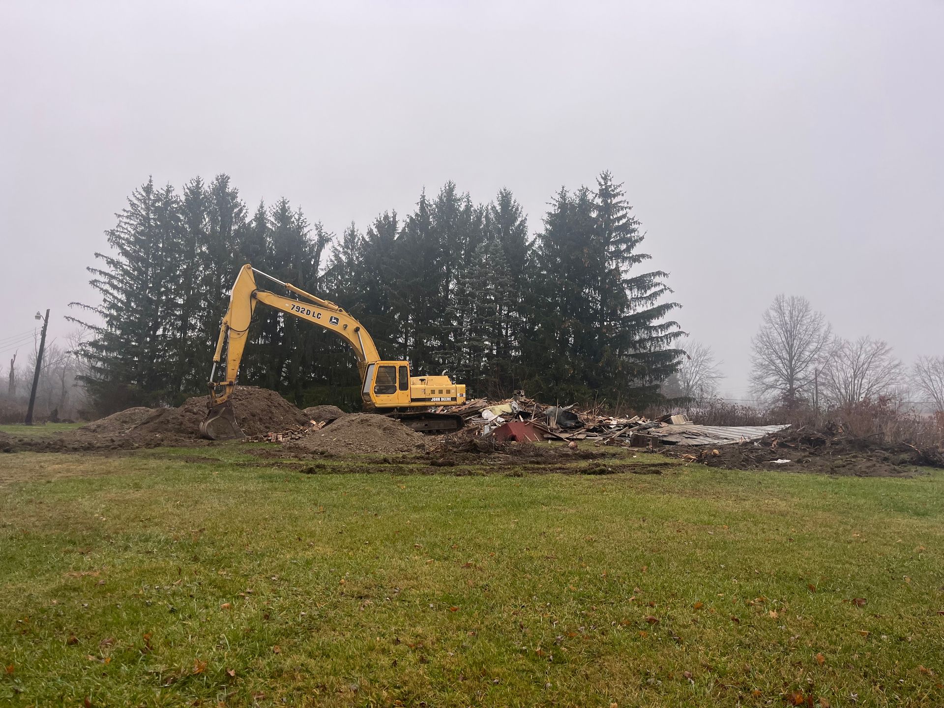 Yellow excavator demolishing structure on a grassy field, trees in the foggy background.