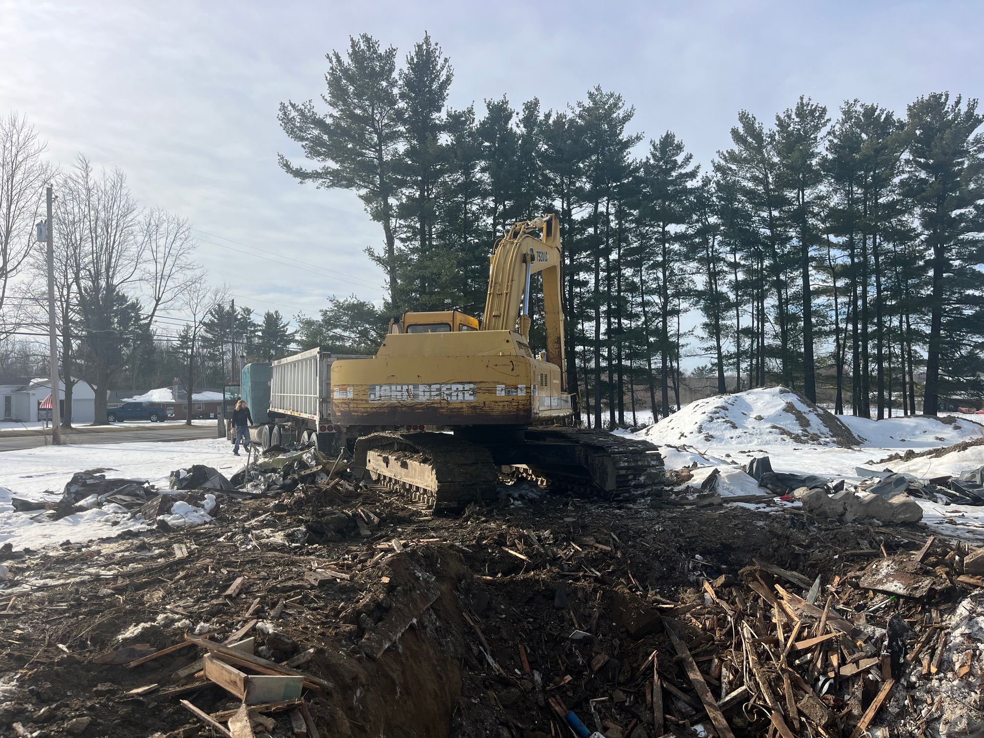 Yellow excavator in a snowy construction site, trees in the background.