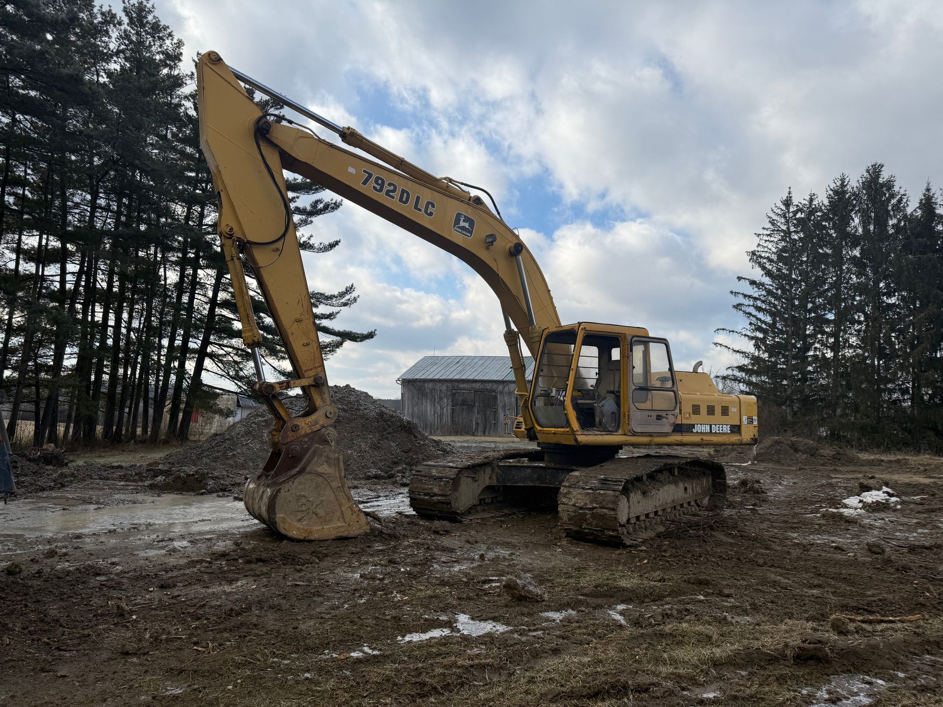 Yellow excavator on muddy ground, near a barn and trees, under a cloudy sky.