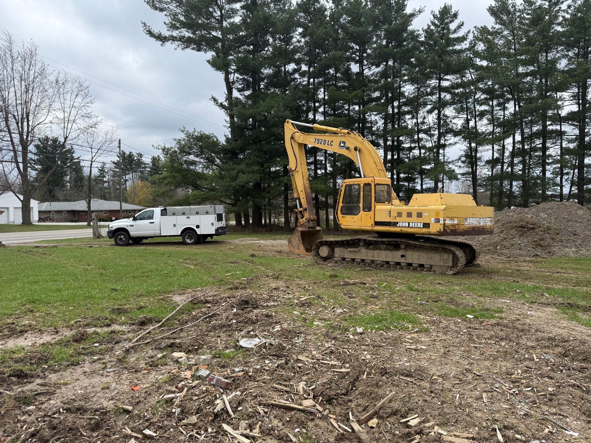 Yellow excavator and white truck on a grassy lot, trees in the background. Construction in progress.
