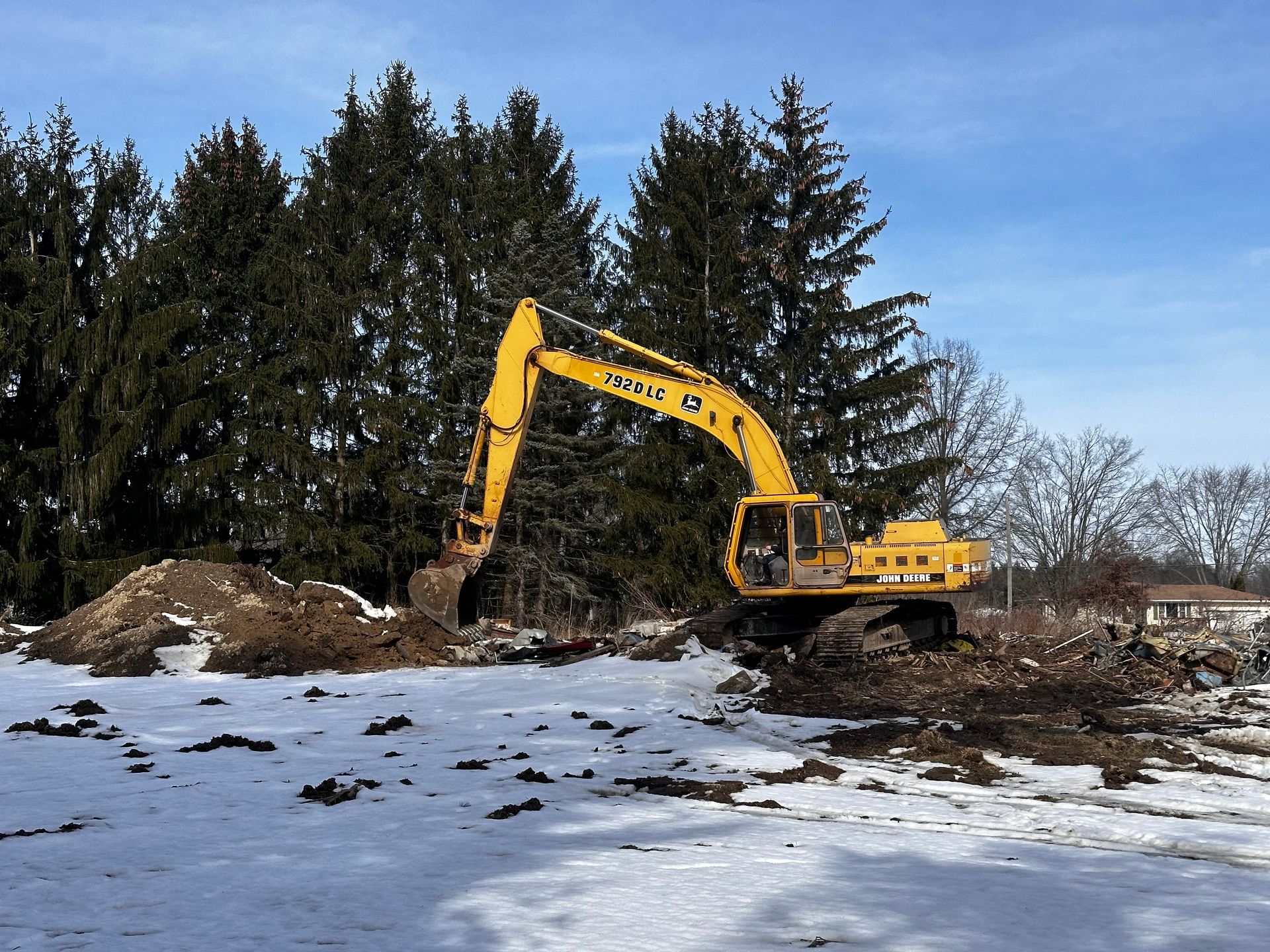Yellow excavator digging in snow, near a pile of dirt and trees against a blue sky.