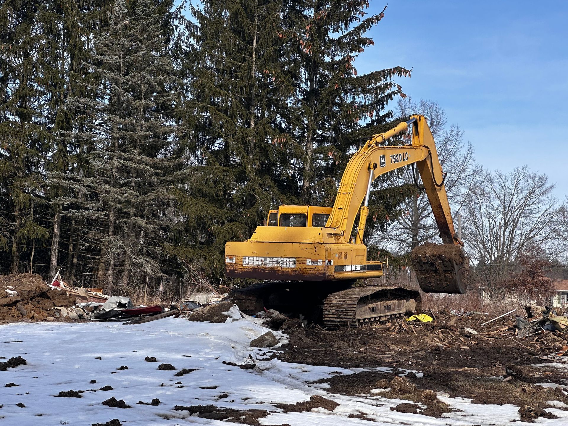 Yellow excavator digging in snow-covered dirt, trees in background, sunny day.