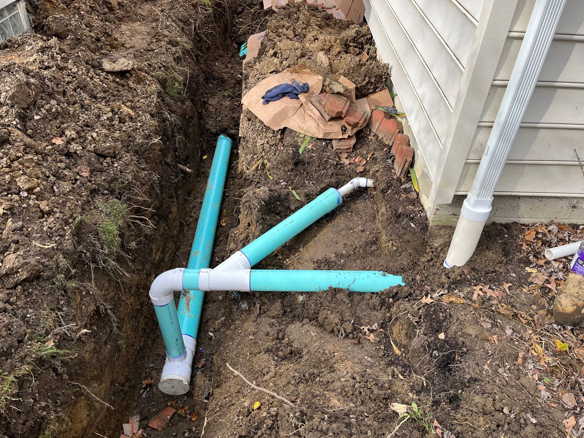 Pipes in a trench near a house's foundation. Turquoise and white pipes with elbows, a downspout visible.
