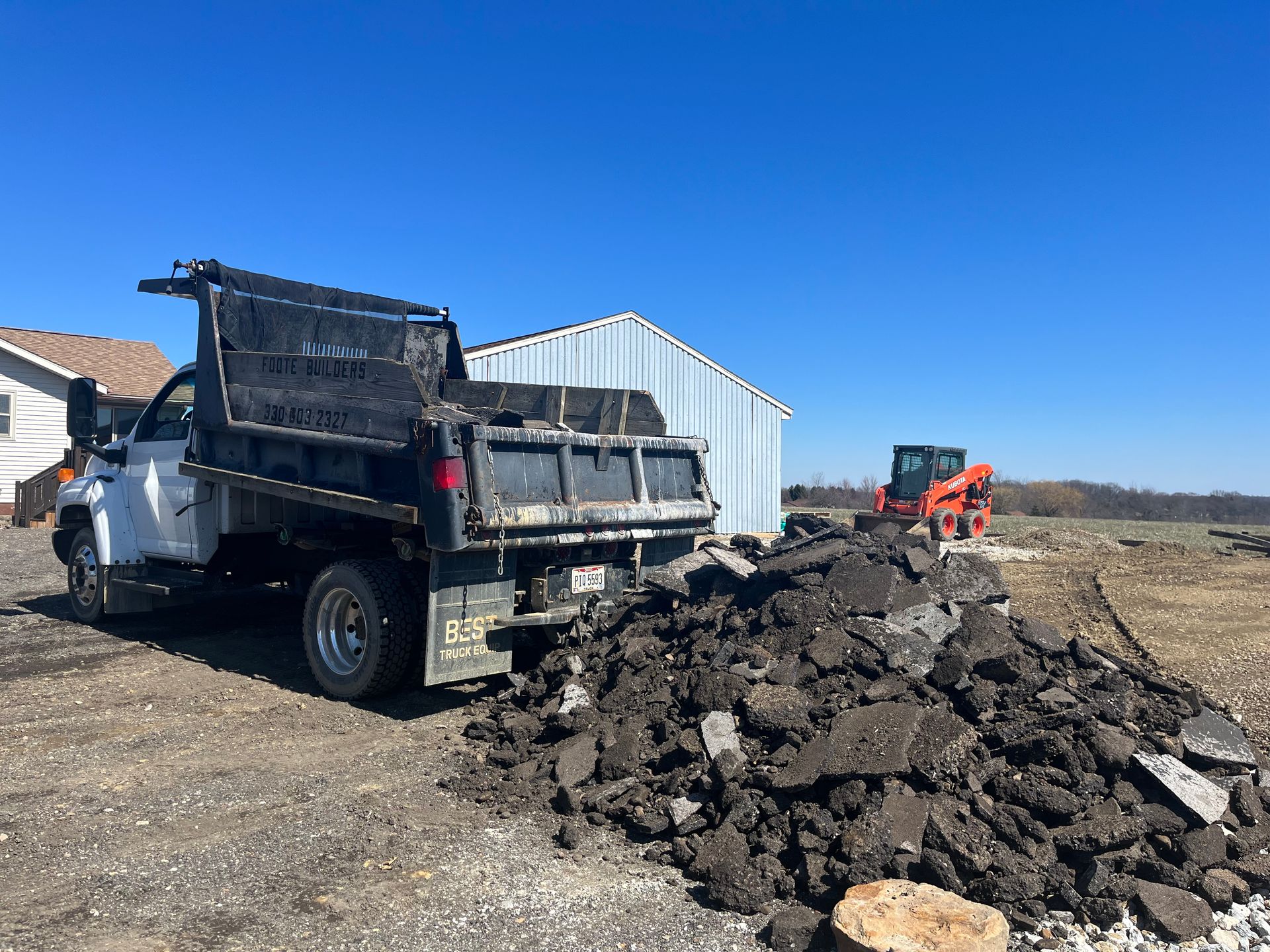 Dump truck unloading asphalt near a shed and small orange skid steer on a sunny day.