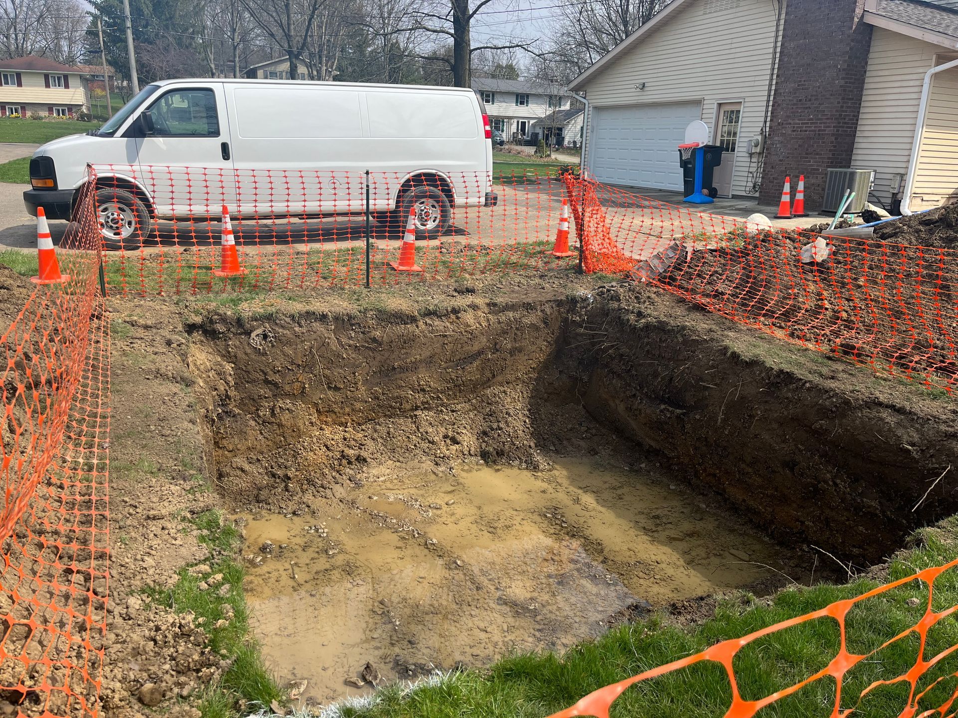 Large excavated area in yard, white van, orange safety fencing, worker near house.