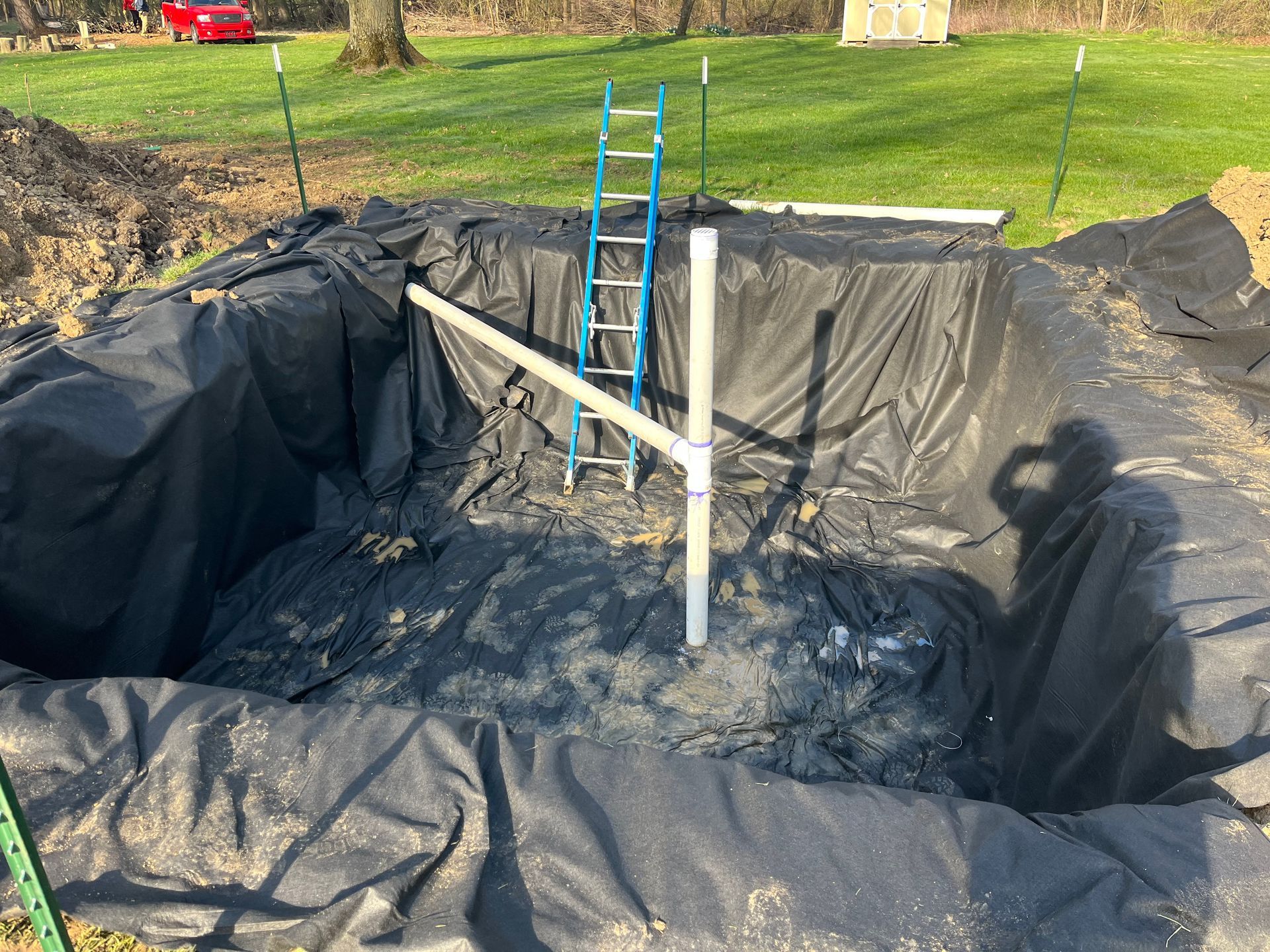 A pond excavation lined with black fabric; a PVC pipe structure and ladder stand inside.