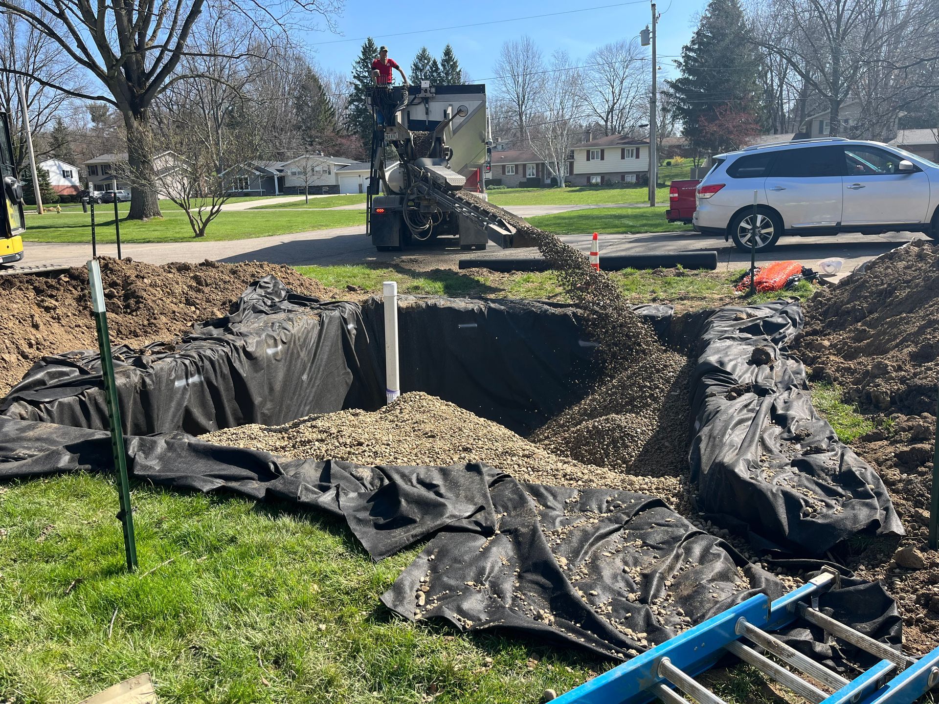 Construction site: gravel being poured into a large hole lined with black fabric; cement truck in the background.