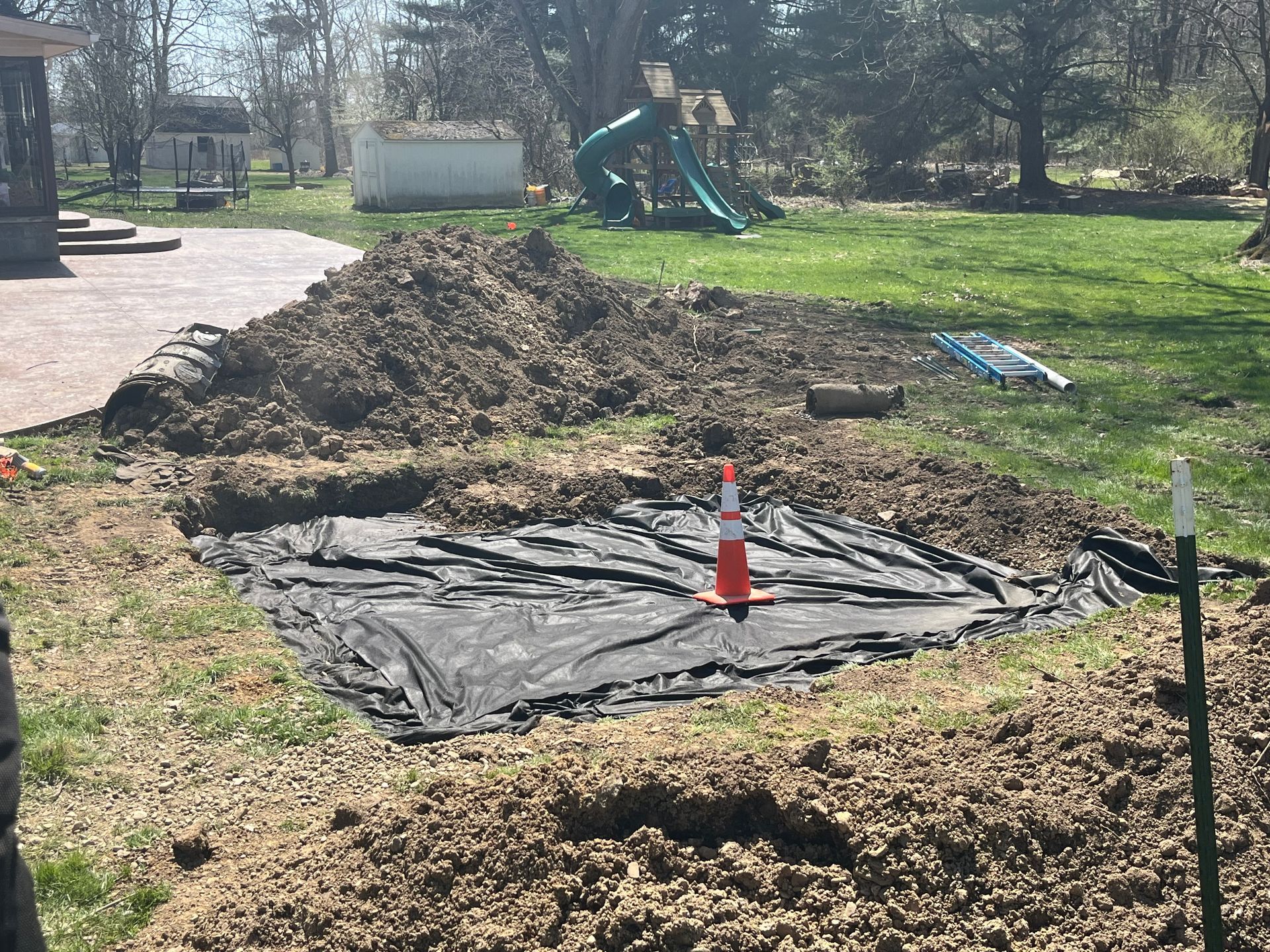 Hole in yard covered with black fabric, dirt pile, traffic cone, green grass, and a swing set in background.