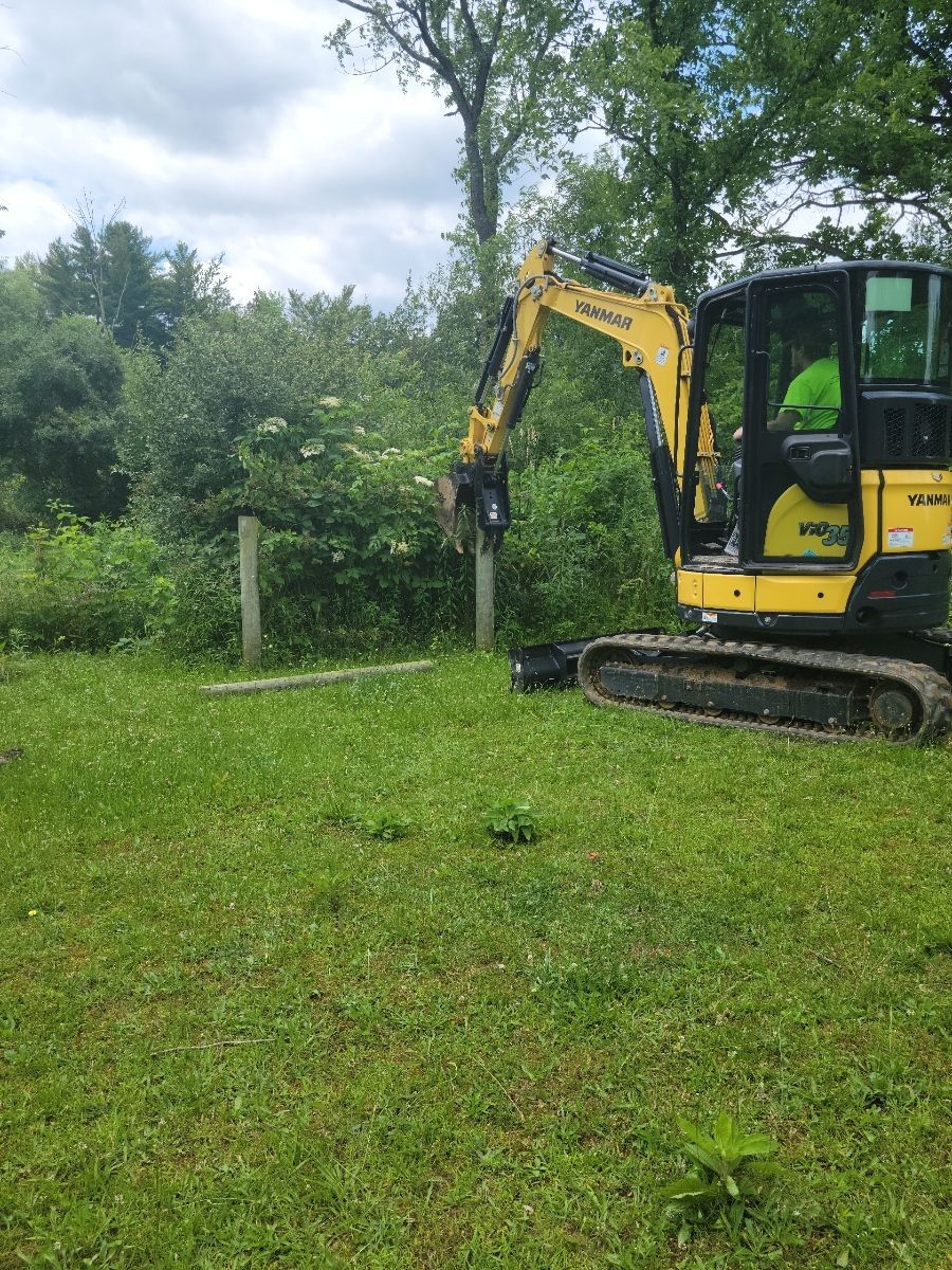 Yellow excavator removing a post in a grassy area with trees and a cloudy sky in the background.