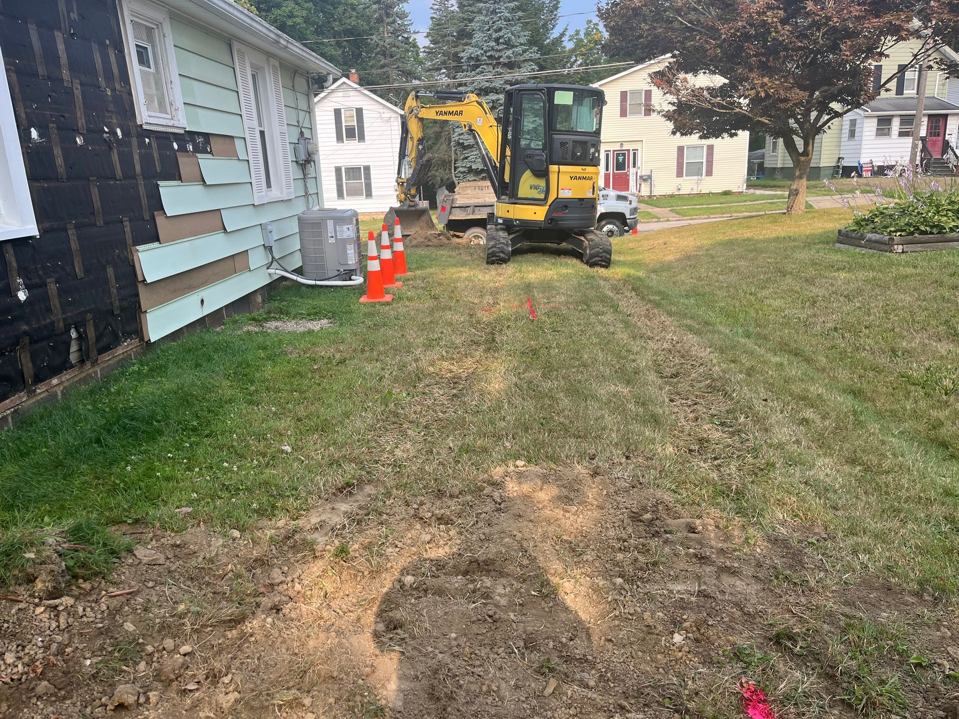 Mini excavator digging near a house, grass and dirt path. Orange traffic cones.