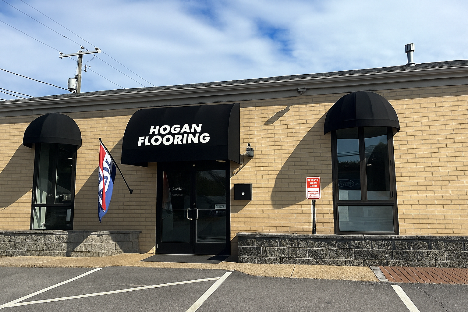 Hogan Flooring storefront with black awnings, windows, and a brick facade. American flag and parking sign.