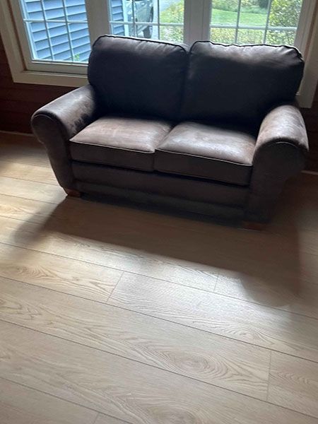 Brown loveseat on light wood floor in front of a window.