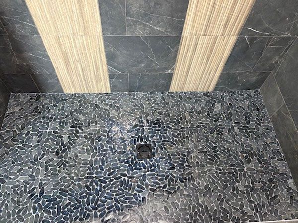 Shower with blue pebble floor, dark tile walls, and light wood-like accent strips.