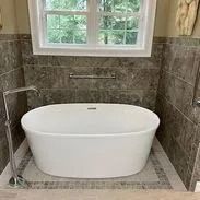 Freestanding white bathtub in a modern bathroom with grey tile walls and a window.