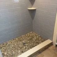 Shower with gray tiled walls and pebble stone floor; corner shelf.