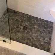 Shower floor with gray pebble tile and drain. White tile wall and glass door.