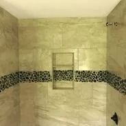 Shower stall with beige tile walls, dark pebble border, and built-in shelf.